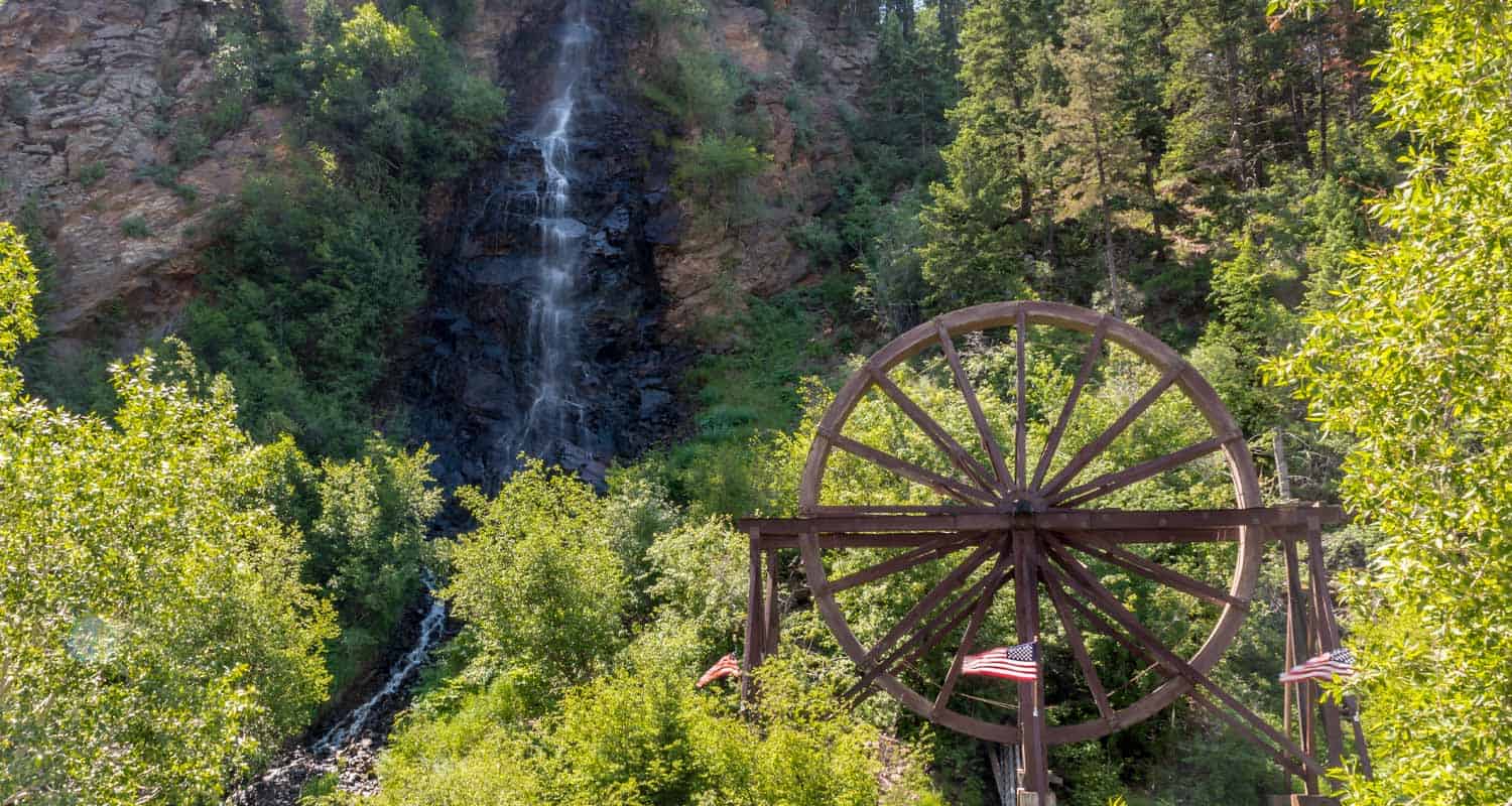 bridal veil falls waterfall pouring over cliff face with waterwheel at base among green trees in idaho springs colorado